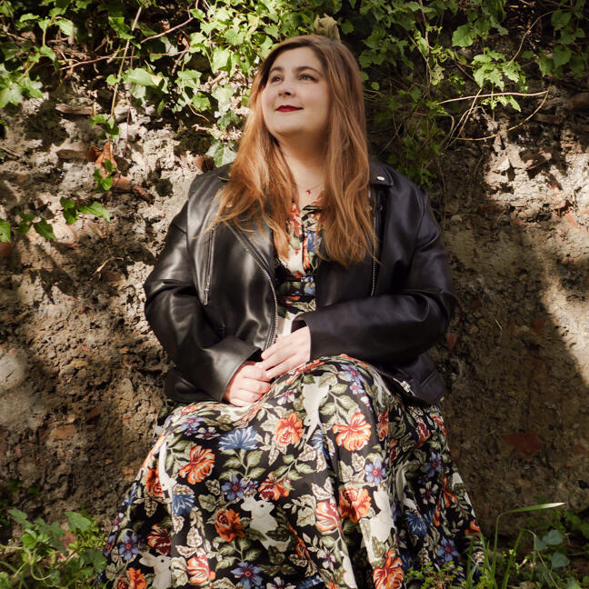 Lizzie, a white person with long ginger-brown hair, sits in front of a brown garden wall covered in ivy.